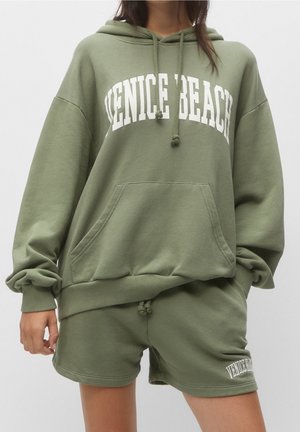 Kobieta nosząca oliwkowo-zielony, oversize'owy kaptur z tekstem "VENICE BEACH" oraz pasujące szorty z małym tekstem "VENICE BEACH" na lewej nodze.