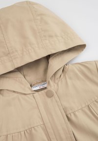 Beżowa kurtka z kapturem wykonana z miękkiego materiału, z zapięciem na napę i subtelnymi detalami szycia. Metka widoczna wewnątrz kaptura.