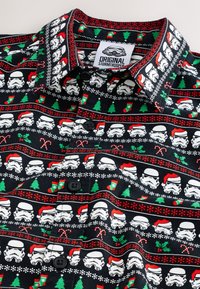 Musta nappipaita, jossa on kuvio, johon kuuluu sotilaita, joulukuusia ja juhlateemaisia punaisen ja vihreän sävyjä mustalla taustalla.