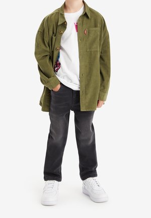 Person trägt ein übergroßes, olivgrünes Hemd über einem weißen T-Shirt mit Grafik, schwarze Jeans und weiße Turnschuhe, steht mit einer Hand in der Tasche.