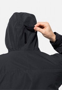 Sort vandtæt jakke med hætte; hånd der griber fat om en Velcro-flap bagpå. Glat struktur, minimalistisk design og ingen synlige logoer.