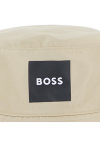 Μπεζ καπέλο τύπου κάσκα από ομαλό ύφασμα, με έντονη μαύρη τετράγωνη ετικέτα που φέρει το λευκό κείμενο "BOSS". Λεπτομερώς ραμμένες άκρες.