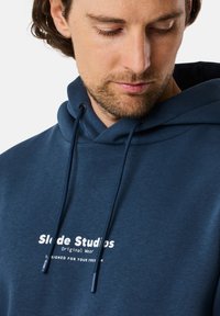 Felpa blu navy realizzata in morbido tessuto, con tasca a marsupio e cappuccio con lacci. La parte anteriore presenta una scritta di marca bianca.