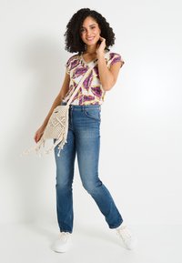 Conjunto casual que incluye un top de punto multicolor con escote en V, jeans de mezclilla azules y un bolso de hombro de crochet en color crema con flecos.