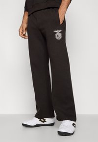 Čierne tepláky vyrobené z bavlnenej zmesi, s elastickým pásom, bočnými vreckami a bielym vyšitým logom na ľavom stehne.