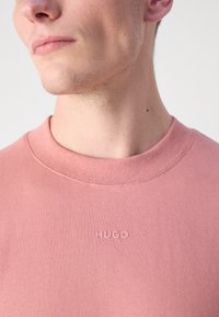Gaiši rozā kokvilnas T-krekls ar noapaļotu dekoltē un diskrētu uzsistā "HUGO" logo uz krūšu daļas. Mīksta tekstūra, ikdienišķa piegriezums.