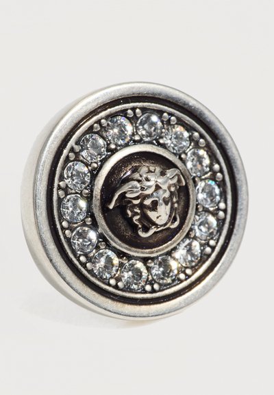 Bouton métallique rond orné d’une tête de Méduse en relief au centre, entourée d’un cercle de pierres précieuses claires et scintillantes.
