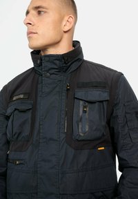 Herre mørkeblå polstret jakke med høj krave, flere lynlås- og klaplommer, tekstureret stof og orange detaljer.