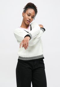 Hvid sweater i kabelstrik med sort-hvide stribede manchetter og kant, med en pink halskant. Kombineret med sorte bukser.