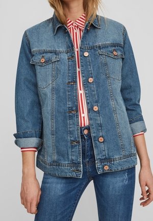 Personne portant une veste en jean bleue par-dessus une chemise à rayures rouges et blanches avec des boutons et un jean bleu, debout devant un fond uni.