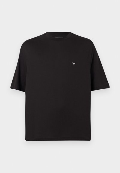 Μαύρο T-shirt με κοντά μανίκια και στρογγυλή λαιμόκοψη, με μικρό λευκό λογότυπο αετού της Emporio Armani στο κέντρο του στήθους πάνω σε γκρι φόντο.