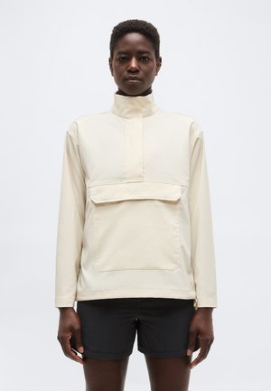 Osoba v krémovom pulóvri s vysokým golierom, veľkou prednou kapsou a čiernymi šortkami stojí pred jednofarebným svetlým pozadím.