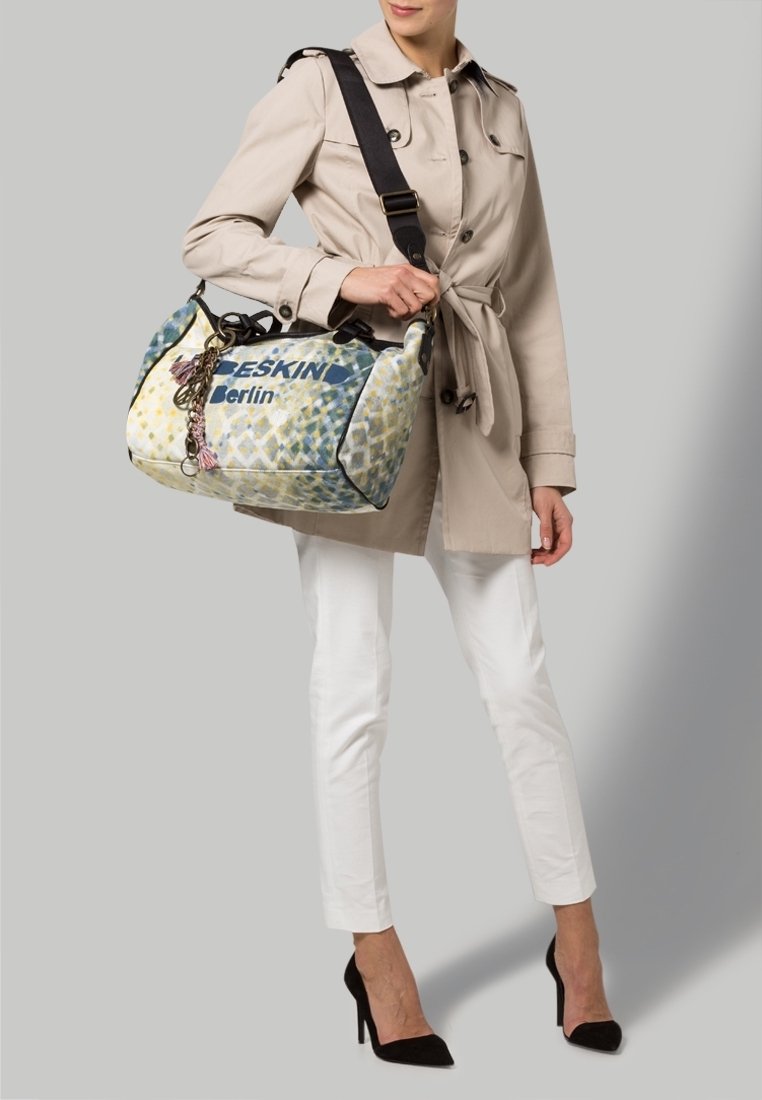 Femme en trench beige et pantalon blanc tenant un sac à motifs Beskin Berlin avec une bandoulière noire, portant des talons noirs.