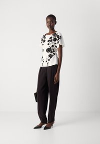 Witte blouse met een zwart bloemmotief, gecombineerd met donkere broeken en zwarte puntige hakken. Accessoires omvatten een kleine zwarte tas.