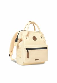 Cream canvas backpack with two top handles, adjustable straps, a front zip pocket, and a round logo patch. Accented with brown hardware.
