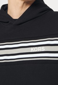 Felpa con cappuccio nera a maniche lunghe con un motivo a righe orizzontali grigie, bianche e nere; presenta il logo "KORS" in bianco sulla riga.