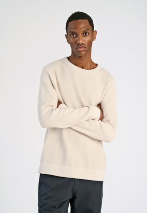 Beige geribbelde trui met lange mouwen, ronde hals en een gestructureerde afwerking, gecombineerd met donkere broeken. Eenvoudig en aansluitend ontwerp.