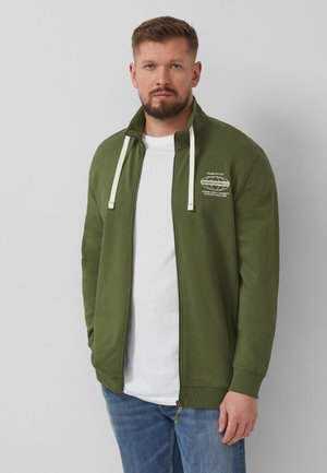 Homme barbu portant une veste zippée verte avec des cordons blancs par-dessus une chemise blanche et un jean bleu, debout devant un fond gris.