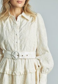 Weißes besticktes Kleid mit kragenförmigem Ausschnitt, langen Puffärmeln und Rüschensaum, in der Taille mit einem weißen Gürtel gerafft.