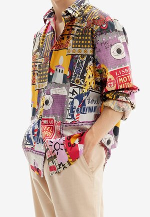 Uomo che indossa una camicia a maniche lunghe colorata e decorata con motivi a blocchi di testo e grafici, abbinata a pantaloni beige, con la mano in tasca.