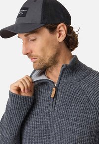 Pull gris à côtes avec un col montant et une fermeture éclair, doté d'un tirette en cuir. Porté avec une casquette noire, montrant un motif en maille texturée.