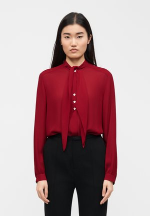 Žena s dlhými čiernymi vlasmi, oblečená v červenej blúzke s mašľou na krku a čiernych nohaviciach, stojaca pred jednoduchým bielym pozadím.