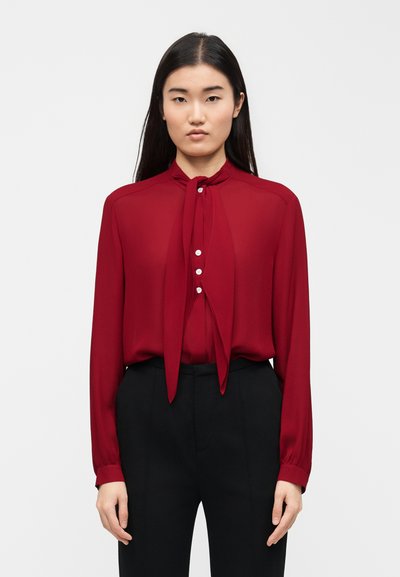 Femme aux longs cheveux noirs portant une blouse rouge avec un nœud à nouer au col et un pantalon noir, debout devant un fond blanc uni.