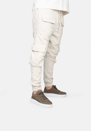 Beige cargo pants with front pockets and ankle cuffs, paired with brown suede sneakers featuring a white sole and lace-up design.