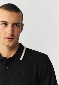 Black polo shirt with beige striped collar, textured fabric, featuring a small embroidered crown logo on the left side.