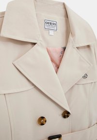Beige trenčkot s velkými klopami, knoflíky ve stylu želvovitého krunýře a růžovou podšívkou s vzorem srdíček a logy. Stříbrné akcenty.