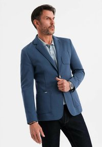 Man justerar en knapp på en blå kavaj, bär en ljusblå polo, mörka jeans, en silverfärgad klocka och armband mot vit bakgrund.