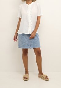 Blouse blanche à boutons avec des détails en volants, associée à une jupe en denim bleu clair avec un motif subtil et des sandales à lanières nouées beiges.