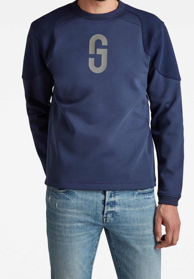 Sweat-shirt bleu marine en tissu doux, avec une grande lettre grise "G" sur le devant et des manches raglan. Porté avec un jean bleu clair.