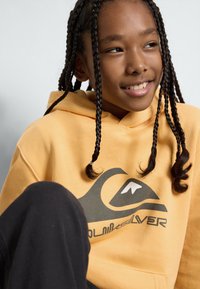 Gul hoodie med en grafisk vågdesign i svart och vitt. Materialet verkar mjukt; den har en rymlig huva och en känguruficka.