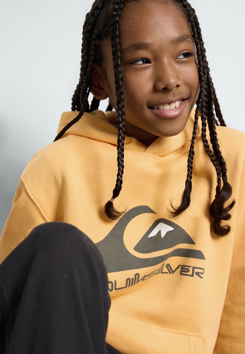 Gul hoodie med en grafisk vågdesign i svart och vitt. Materialet verkar mjukt; den har en rymlig huva och en känguruficka.