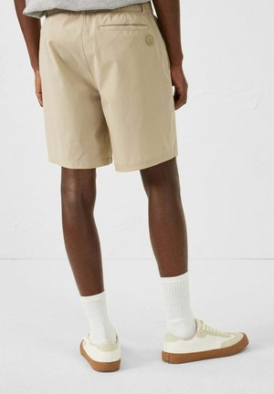 Pantalones cortos beige con cinturilla elástica y bolsillo trasero, combinados con zapatillas crema y calcetines blancos. Logo visible en los pantalones cortos.