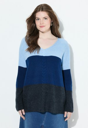Giovane donna con lunghi capelli castani che indossa un maglione morbido a blocchi di colore in azzurro chiaro, blu navy e grigio scuro, in piedi davanti a uno sfondo bianco.