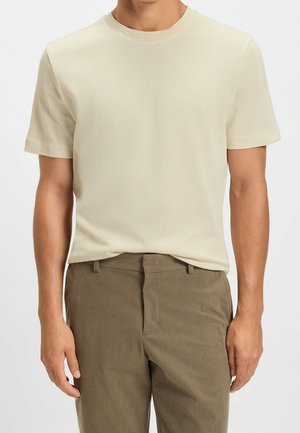 Man draagt een effen lichtbeige kort mouw gebreid shirt, ingestopt in lichtbruine broek met hoge taille, tegen een witte achtergrond.