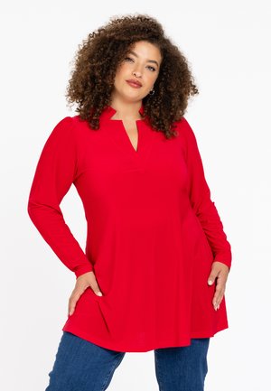 Rode tuniek met lange mouwen en een gespleten halslijn, relaxte pasvorm en zachte textuur. Gecombineerd met blauwe jeans, voorzien van subtiele plooien bij de mouwen.