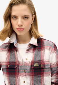 Chemise en flanelle à carreaux rouge, gris et blanc, avec un bouton devant, un col et une poche poitrine arborant un écusson logo jaune et noir.