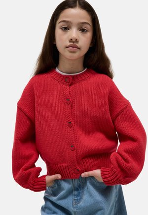 Jeune personne aux longs cheveux porte un cardigan rouge tricoté oversized et un jean bleu, mains dans les poches, expression neutre.