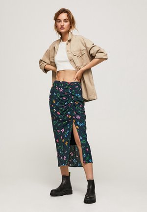 Femme portant une veste beige, un top court blanc, une jupe midi noire à fleurs avec une fente à l'avant, et des bottines noires, debout devant un fond uni.