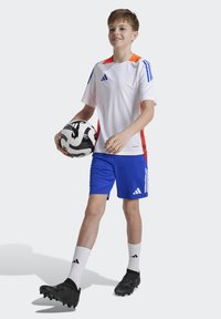 Camiseta atlética blanca con acentos en naranja y azul, combinada con pantalones cortos azules. Zapatillas de fútbol negras y calcetines blancos con rayas negras. Sosteniendo un balón de fútbol en blanco y negro.
