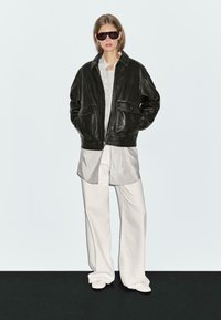 Giubbotto bomber in pelle nera con due tasche frontali, camicia a righe bianche, pantaloni bianchi a gamba larga e occhiali da sole scuri. Sfondo neutro.