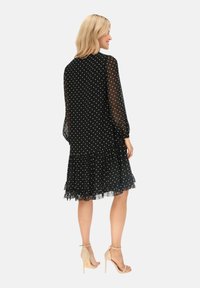Czarna sukienka w kropki z długimi, przezroczystymi rękawami, falbaną u dołu i luźnym krojem. Szczegóły obejmują falbany na dole.