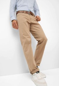 Beige katoenen broeken met een rechte pasvorm, voorzien van een bruine riem. De stof heeft een gladde textuur, met opgerolde mouwen die witte sneakers onthullen.