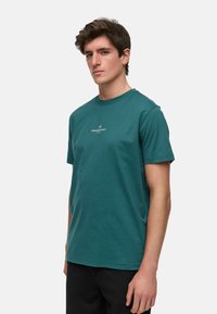 Teal bomull T-skjorte med korte ermer og rund hals, med en hvit "KRONSTADT"-logo på brystet. Minimalistisk design.