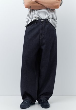 Osoba nosiaca široké čierne rifle, pruhované krátke rukávy vložené dovnútra a čierne nazúvacie topánky, stojaca so založenými rukami.