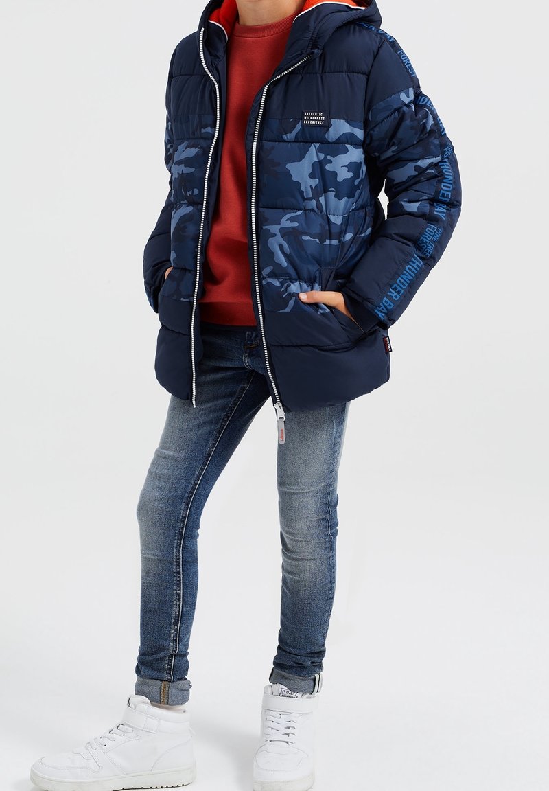 Kind trägt eine blaue Camouflage-Daunenjacke, einen roten Pullover, ausgewaschene Skinny-Jeans und weiße High-Top-Sneaker, steht mit den Händen in den Taschen.