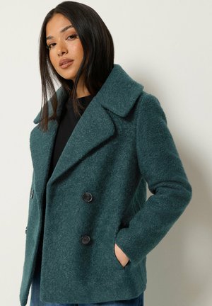 Femme aux cheveux raides et foncés portant un manteau en laine bleu sarcelle à double boutonnage, les mains dans les poches, sur un fond clair uni.